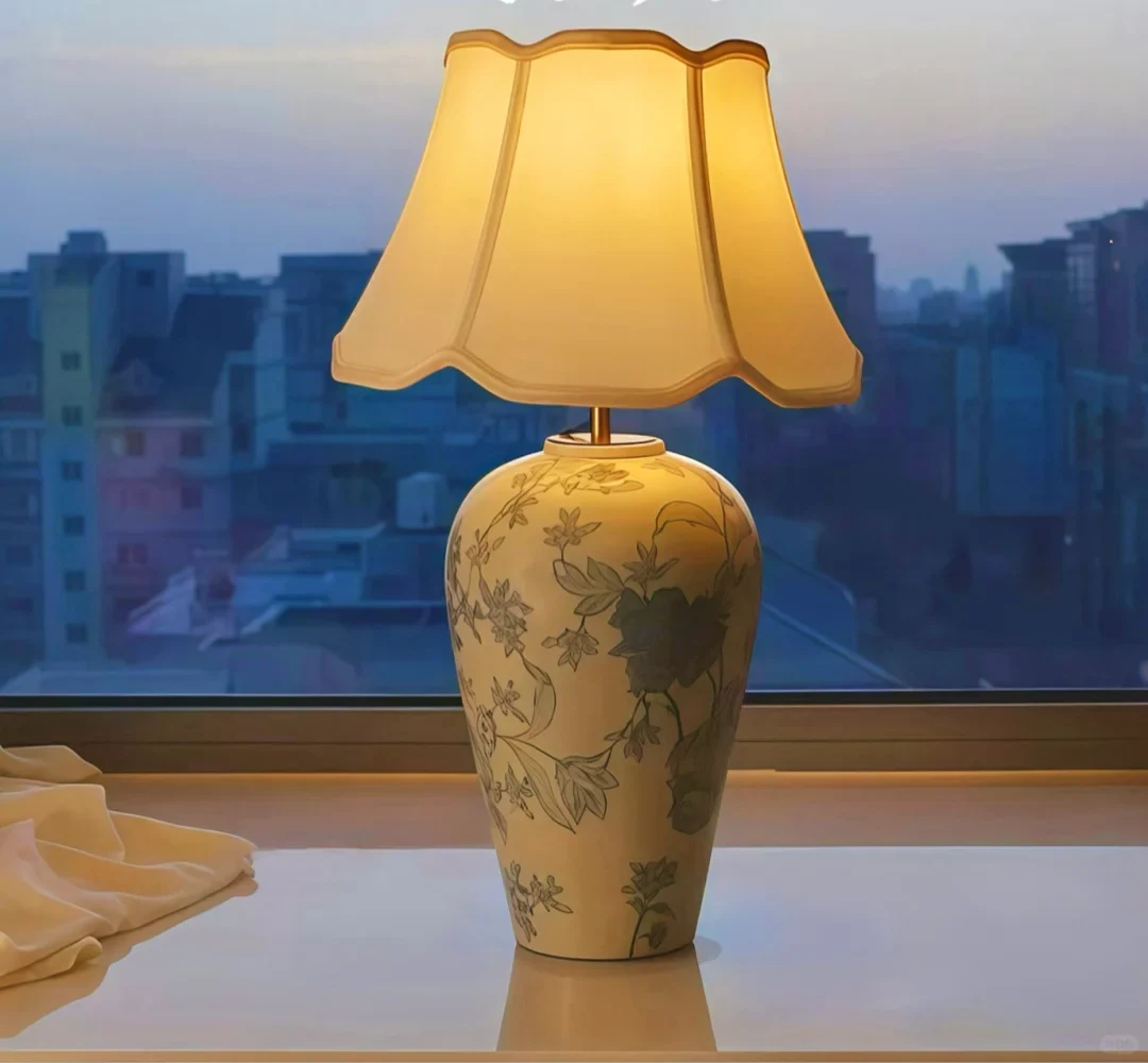 A floral ceramic vase lamp with a warm scalloped shade against the blue evening outside a window
