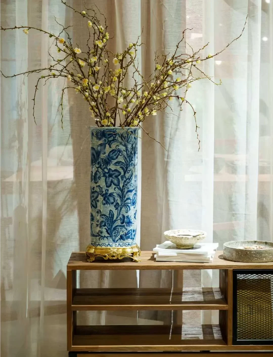 Blue-and-white cylindrical vessel on a gilt-colored mount holding flowering branches beside a window