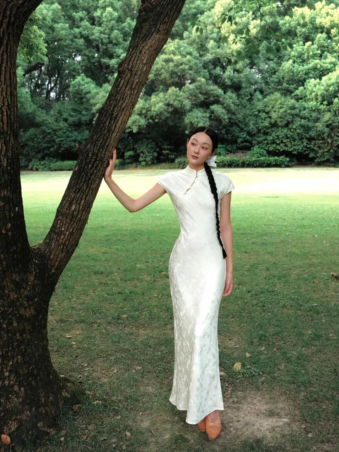 A woman wearing a full-length white qipao in Shanghai Guilin Park, with the standing collar, fitted waist and narrow hem clearly visible