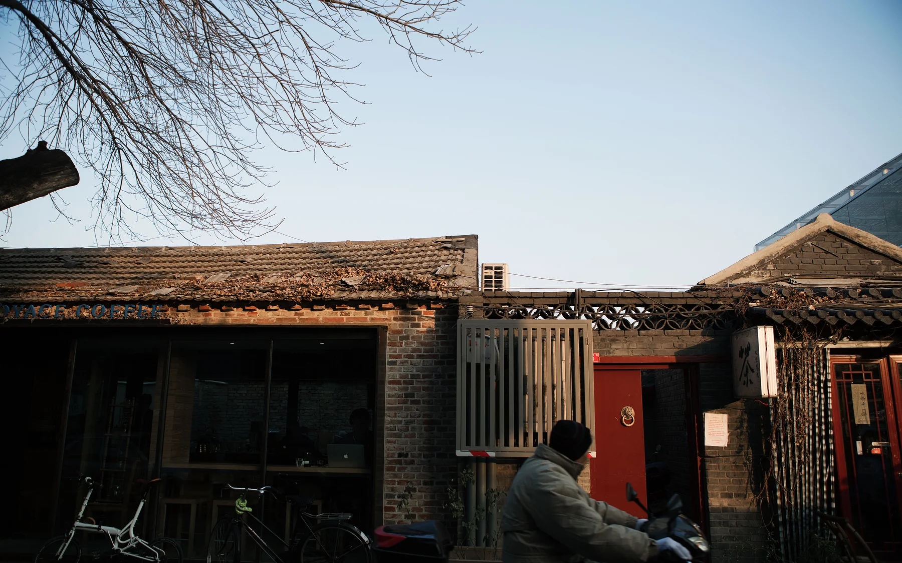 A rider passing old courtyard roofs and a small cafe in a Beijing hutong, showing ordinary city rhythm beyond landmark hours