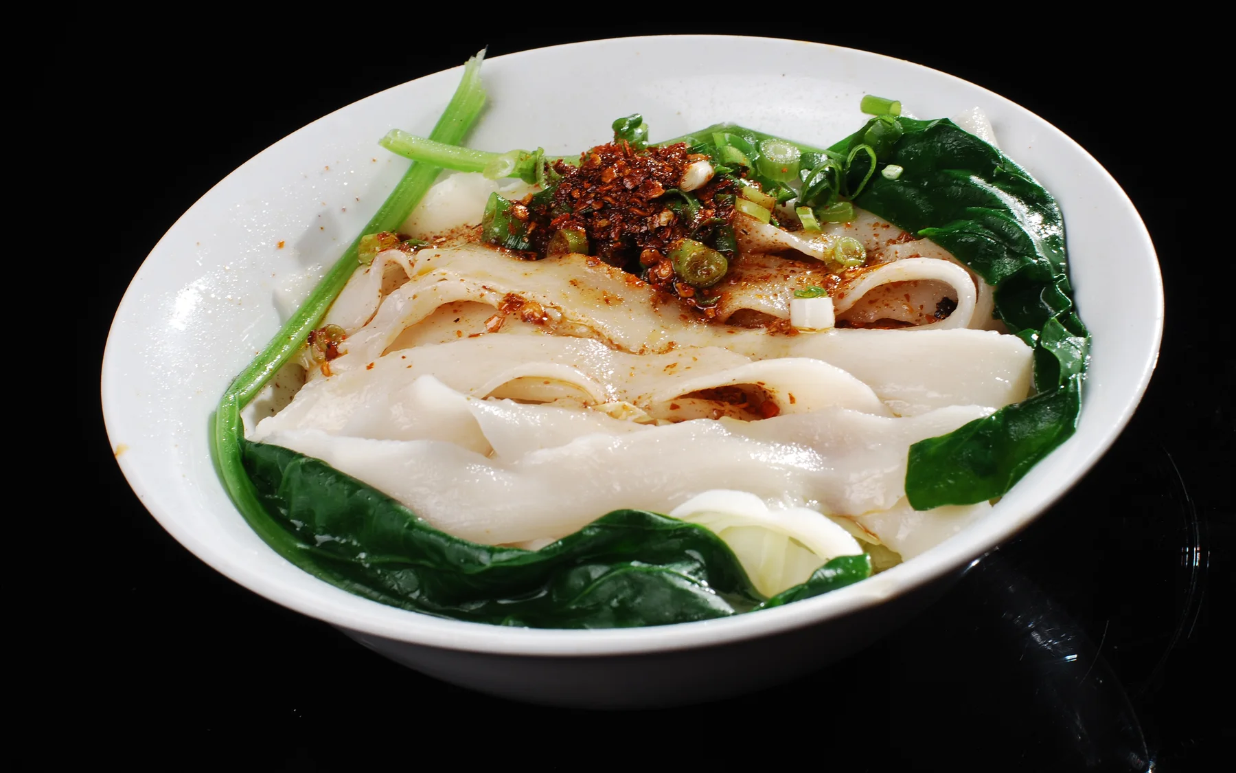 A broad bowl of oil-splashed noodles showing wheat texture, chile and scallion at the table