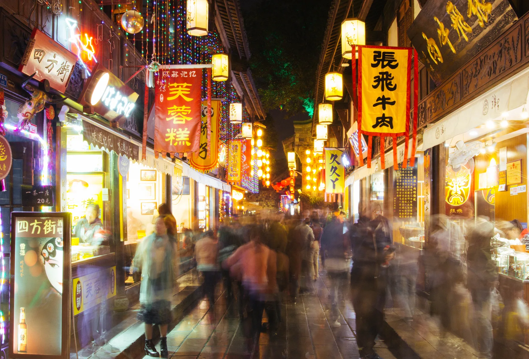 People move between bright food signs on a crowded Chengdu street at night