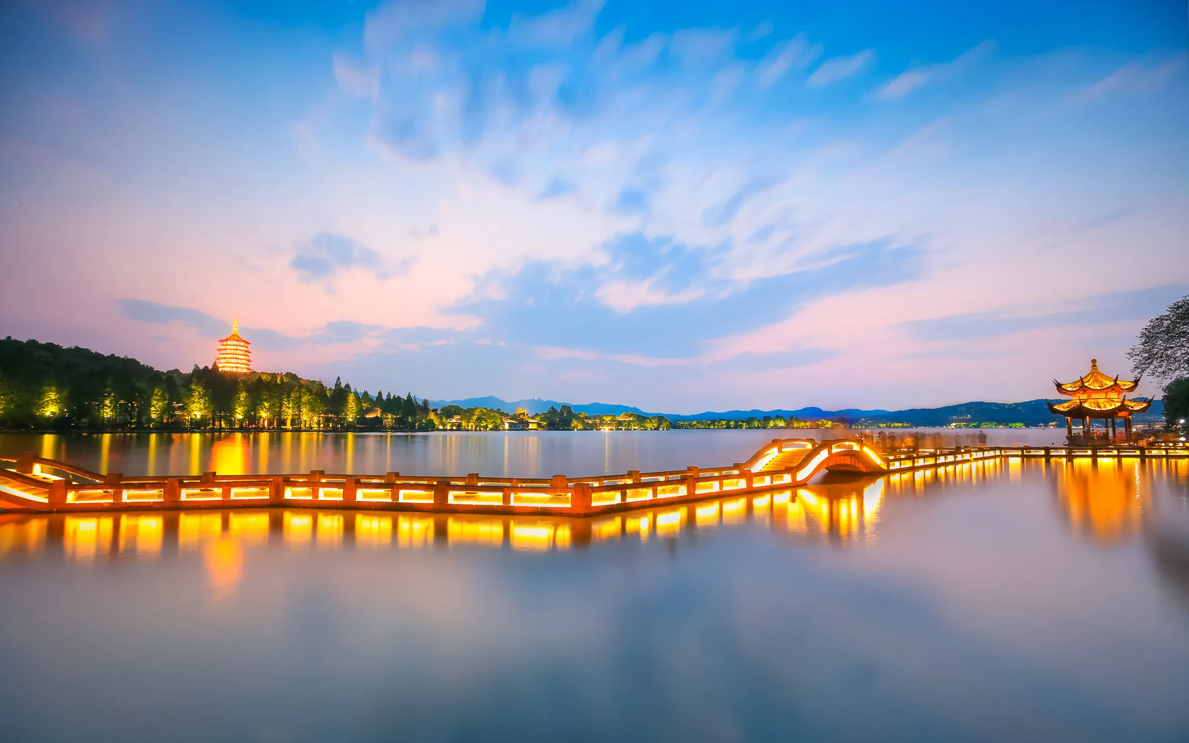 West Lake in Hangzhou at blue hour with Leifeng Pagoda, a lit causeway, pavilion and wooded hills