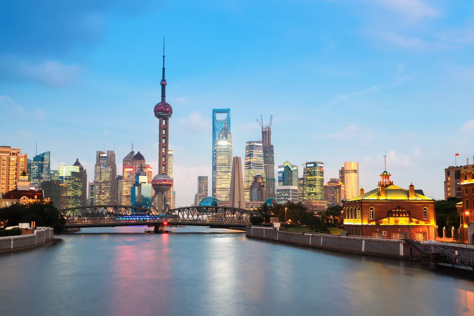 Historic riverside buildings, an iron bridge and the Pudong skyline layered across a Shanghai waterway at dusk