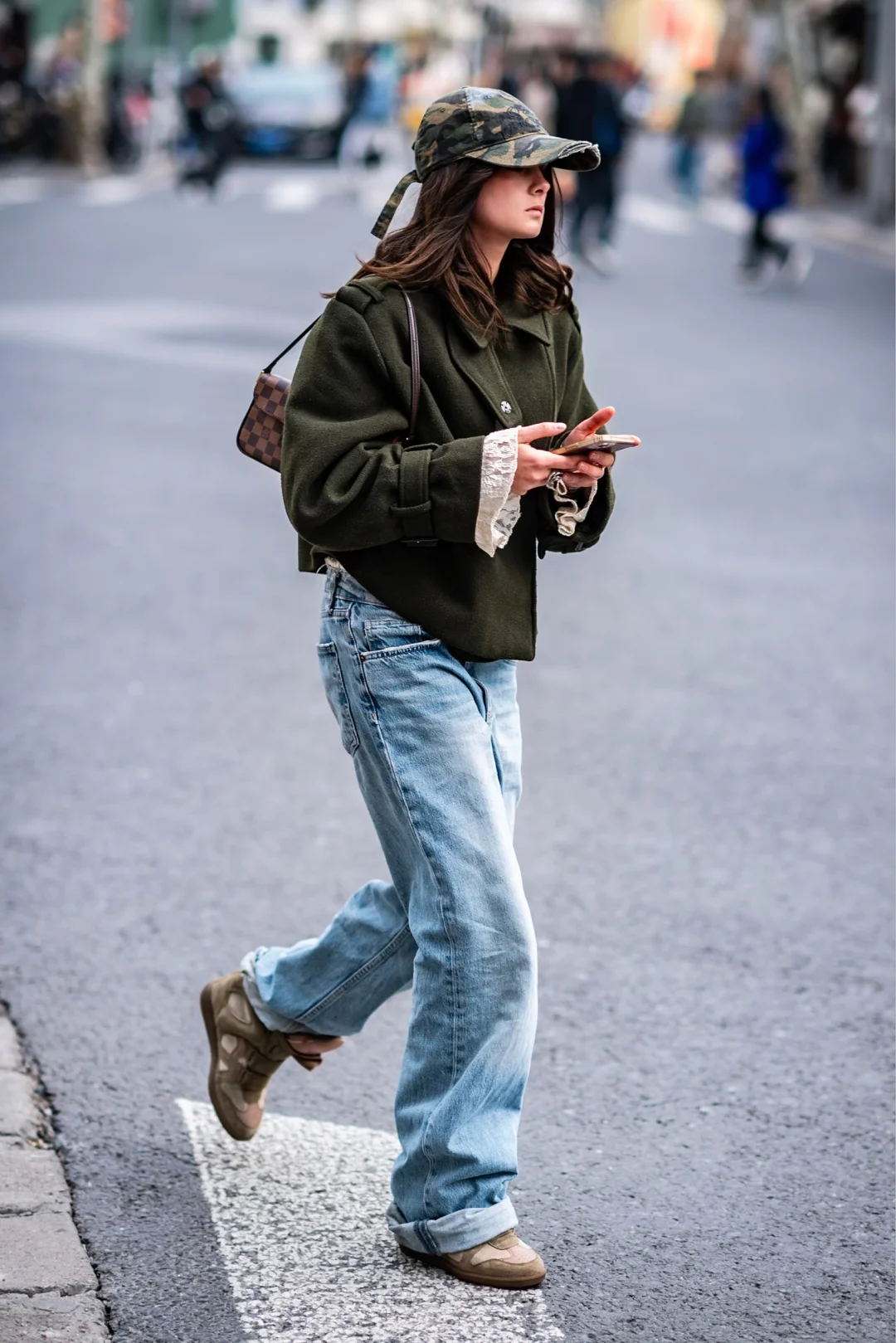 A pedestrian in a camouflage cap, cropped green coat, lace cuffs and wide faded jeans checking a phone while crossing