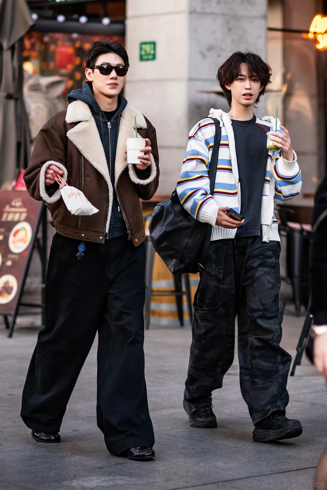 Two people walking with drinks in layered shearling, hoodies, striped knit and dark wide trousers