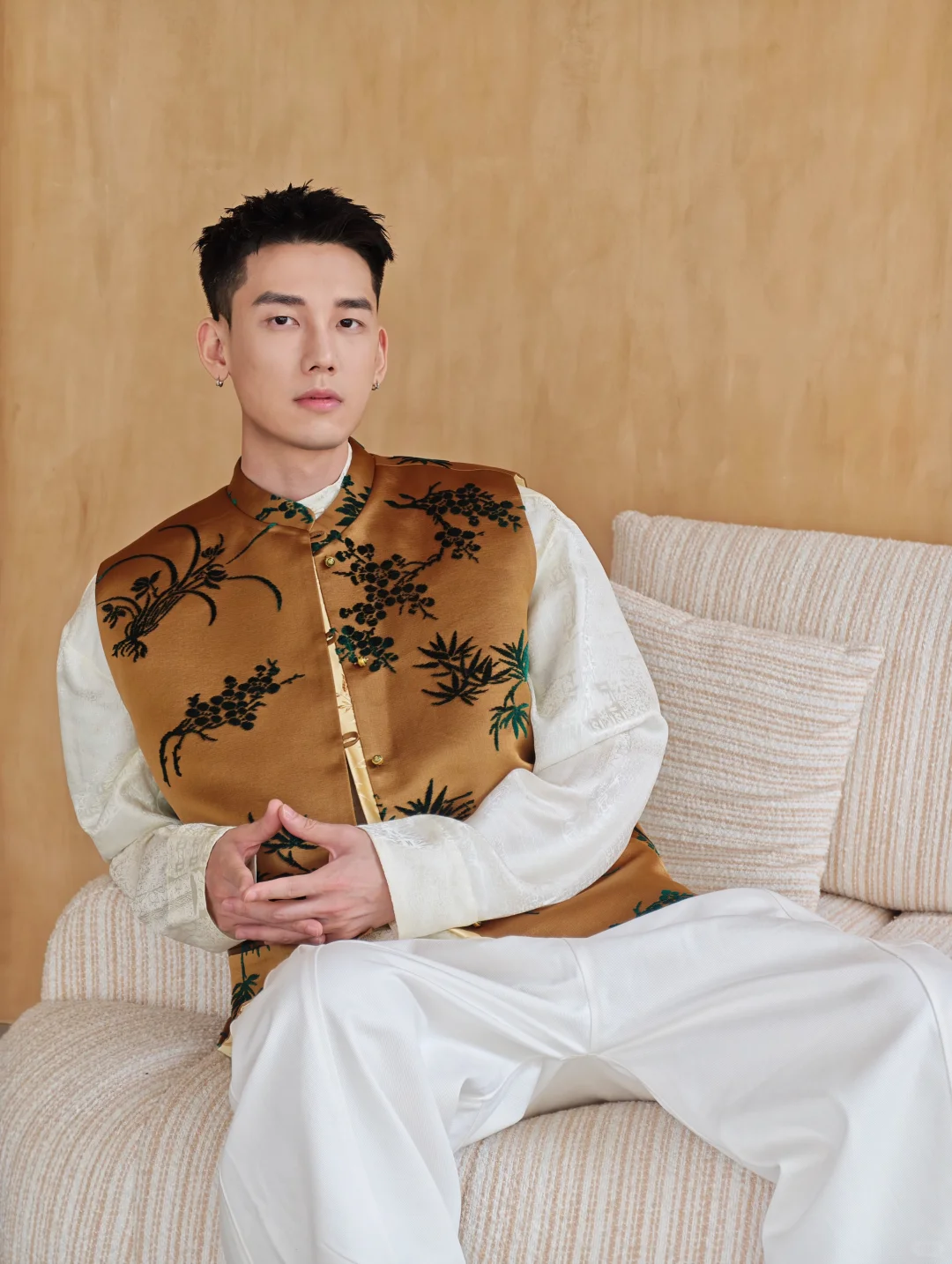 A seated model wearing a brown-gold botanical vest over an ivory patterned shirt and wide white trousers
