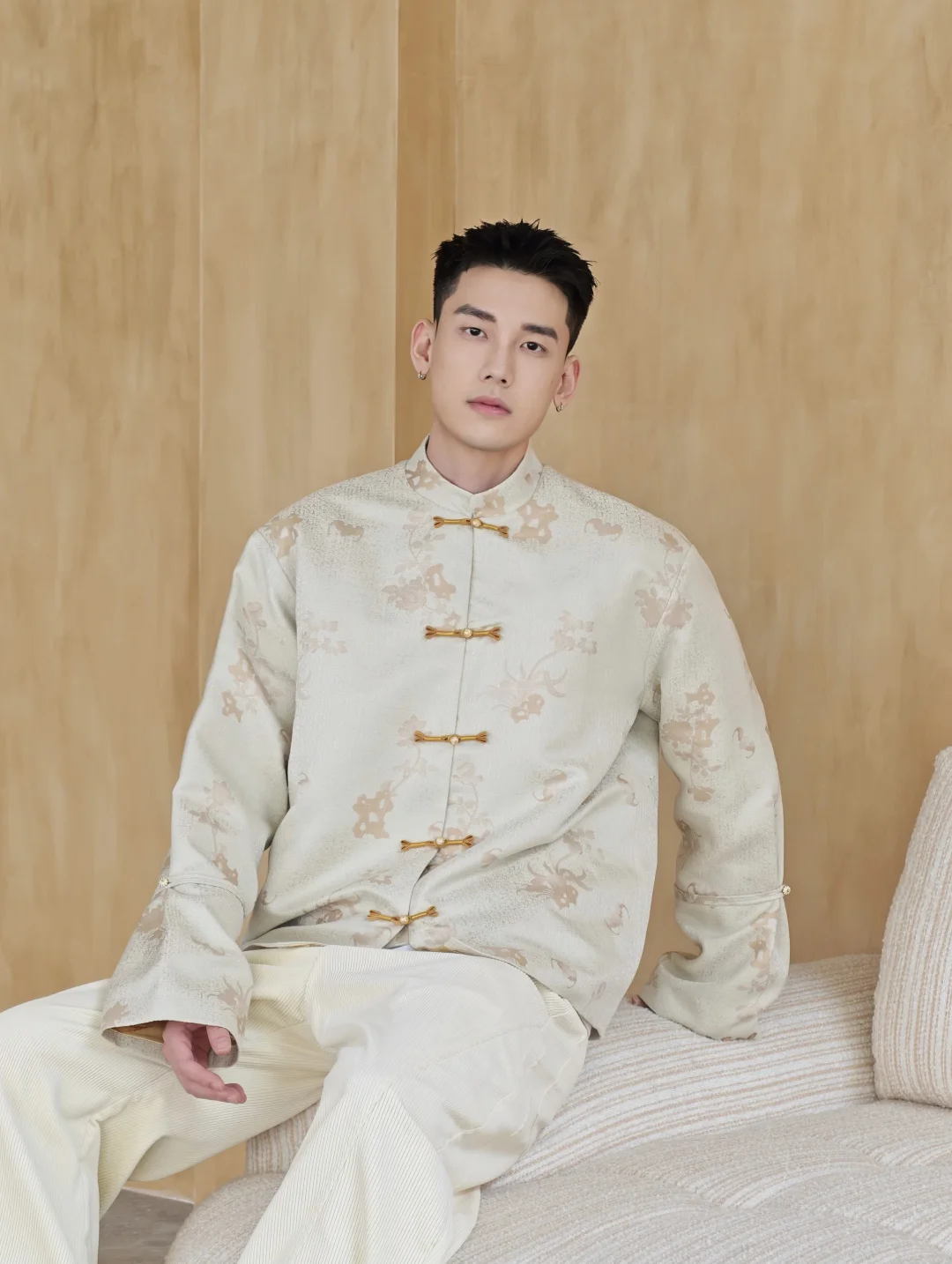 A model seated in an ivory jacket with five gold-toned knot fastenings and softly patterned cloth