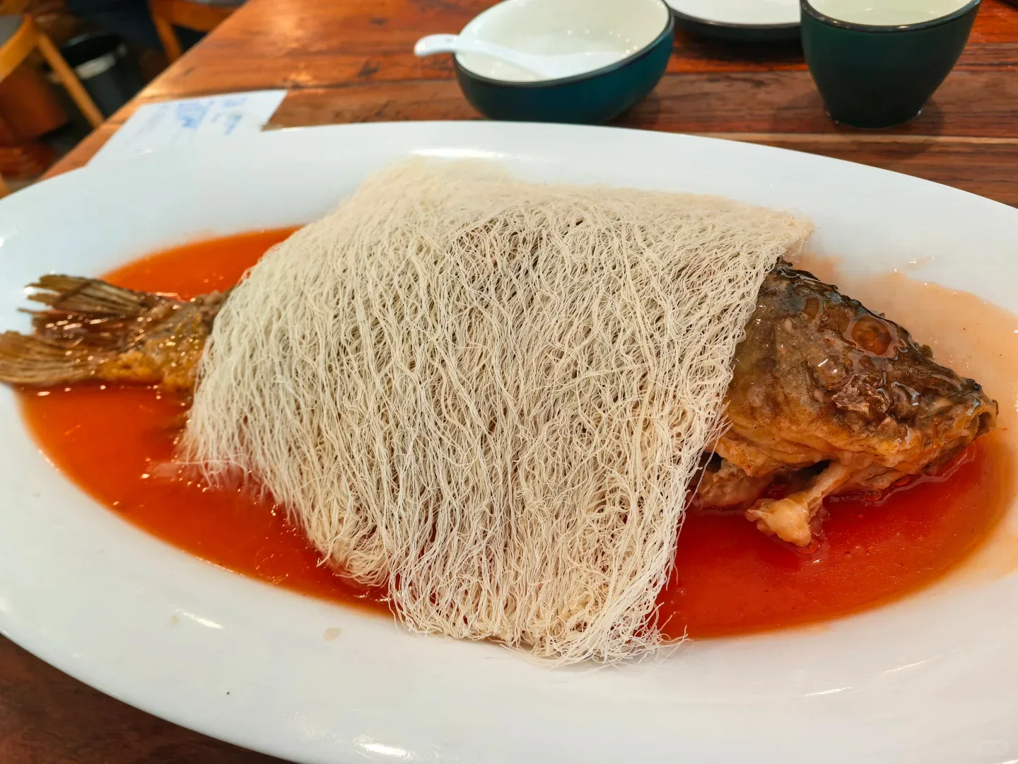 Whole Kaifeng carp on an oval platter, head and tail visible around a pale sheet of fine fried noodles and red sweet-sour sauce