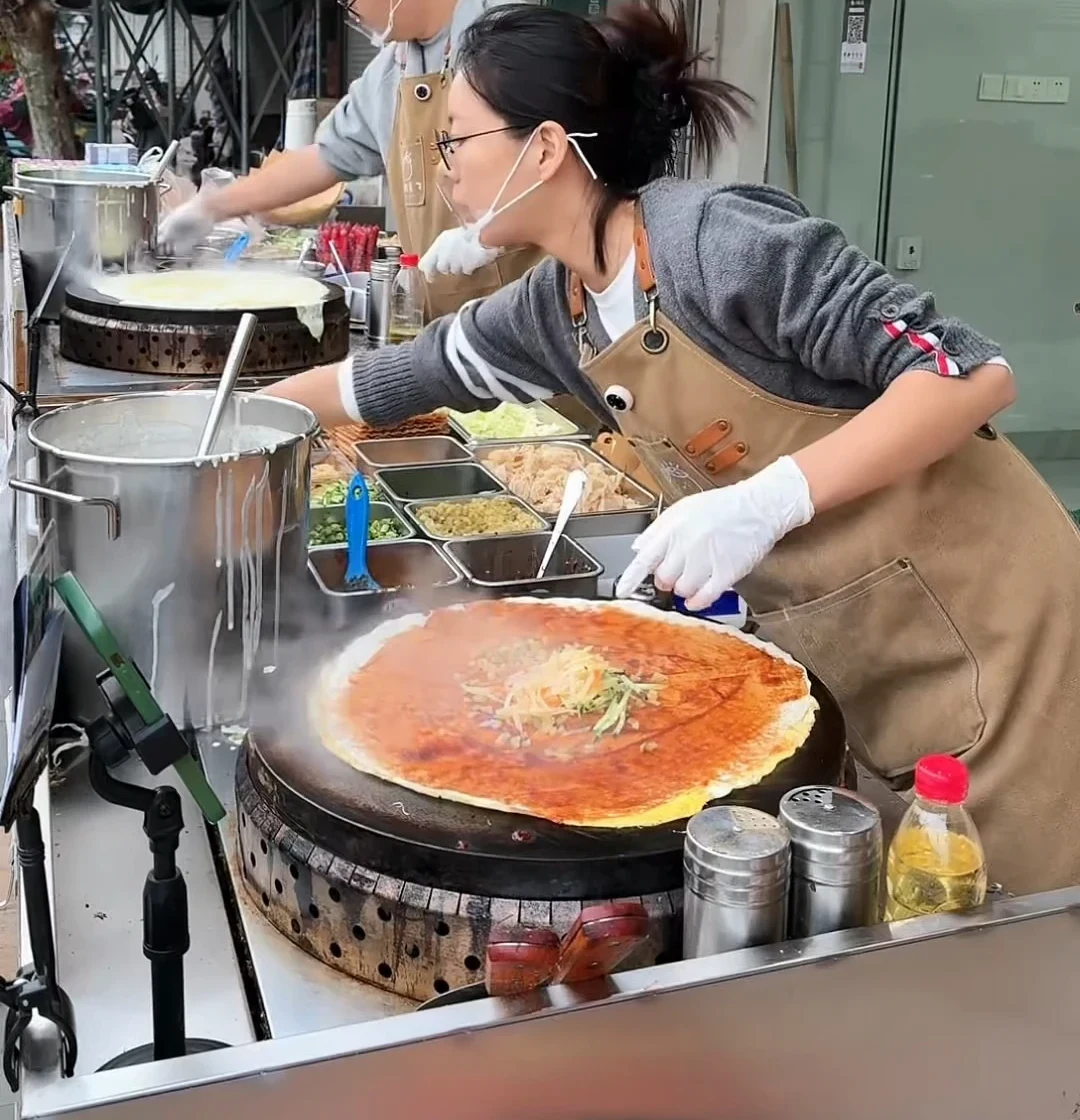 Two breakfast-stall workers assembling jianbing simultaneously across neighboring griddles