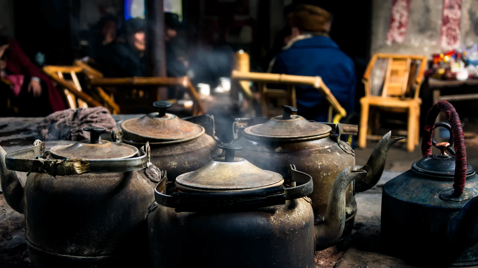 Well-used metal kettles steaming at an old Chengdu teahouse while guests sit behind them