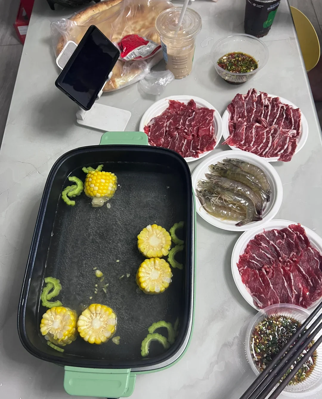 A home Chinese hot pot table with clear broth, thinly sliced beef, shrimp, corn, bitter melon and two dipping sauces