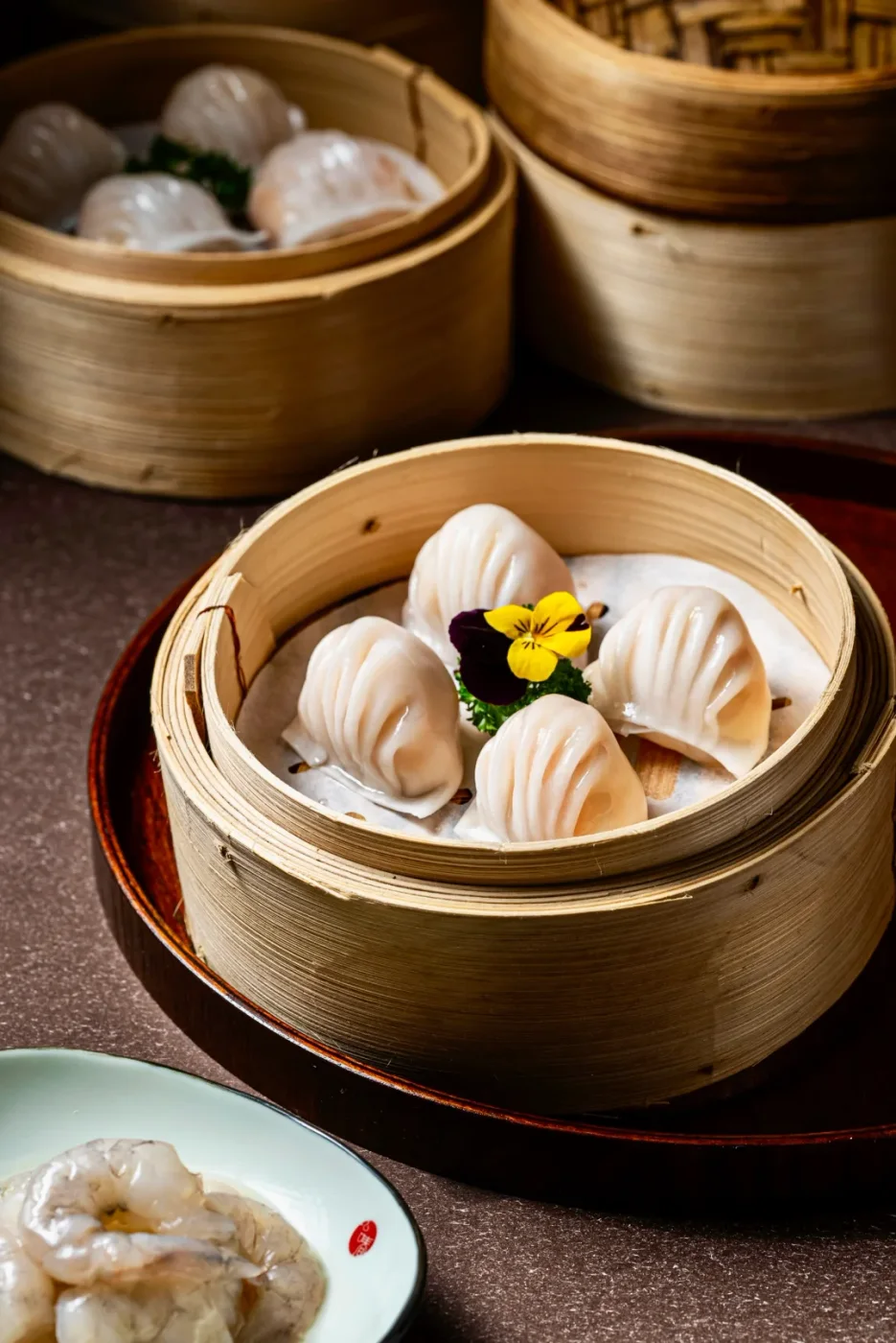 A bamboo steamer holds four translucent har gow beside other dishes on a real yum-cha table