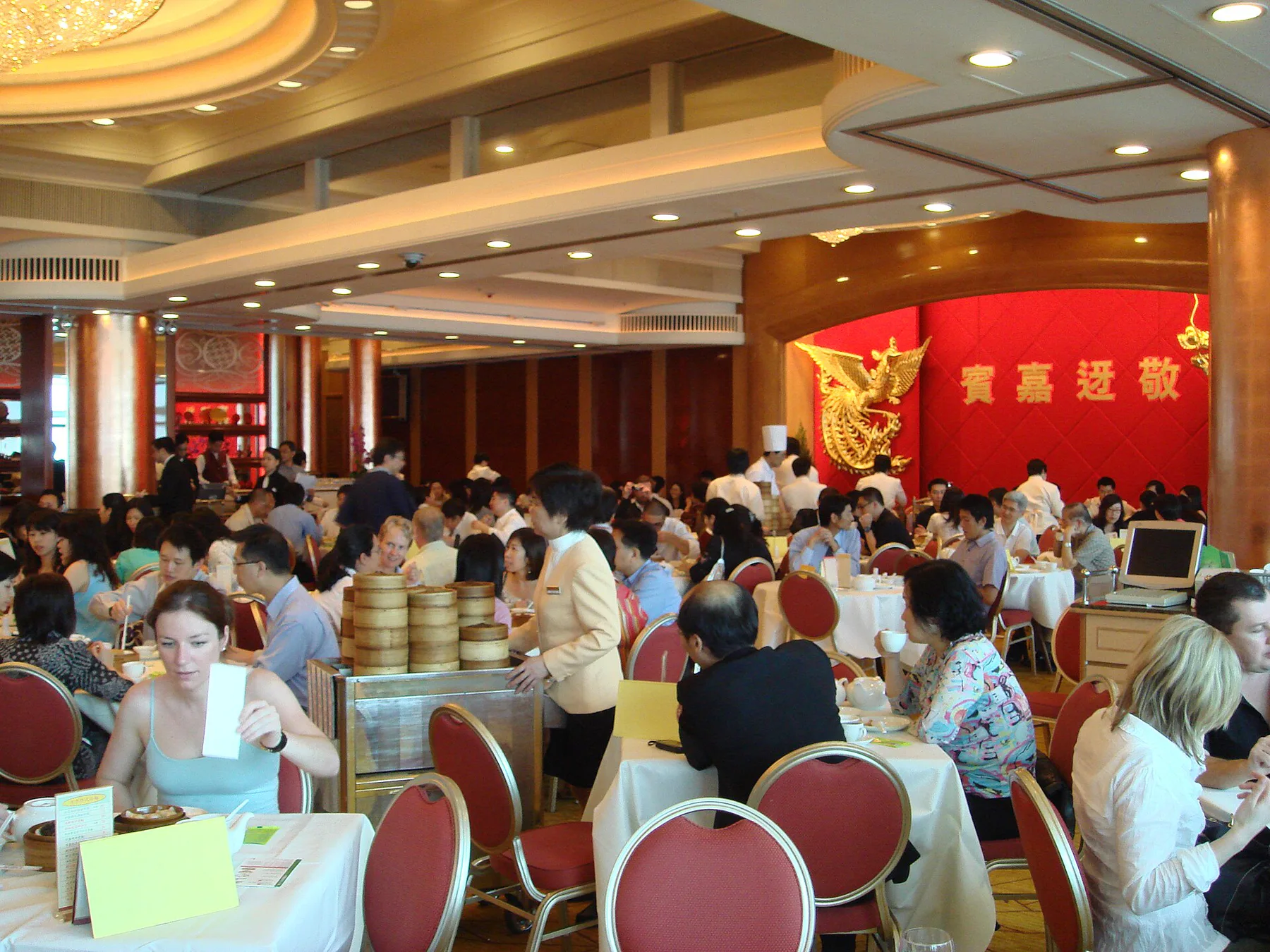 A busy Hong Kong yum-cha dining room with diners sharing round tables while staff move a cart stacked with bamboo steamers