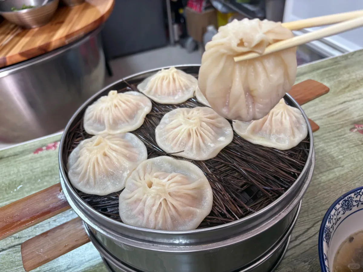 Real Kaifeng street-side basket of pale pleated soup dumplings, one intact dumpling held above the steamer with chopsticks