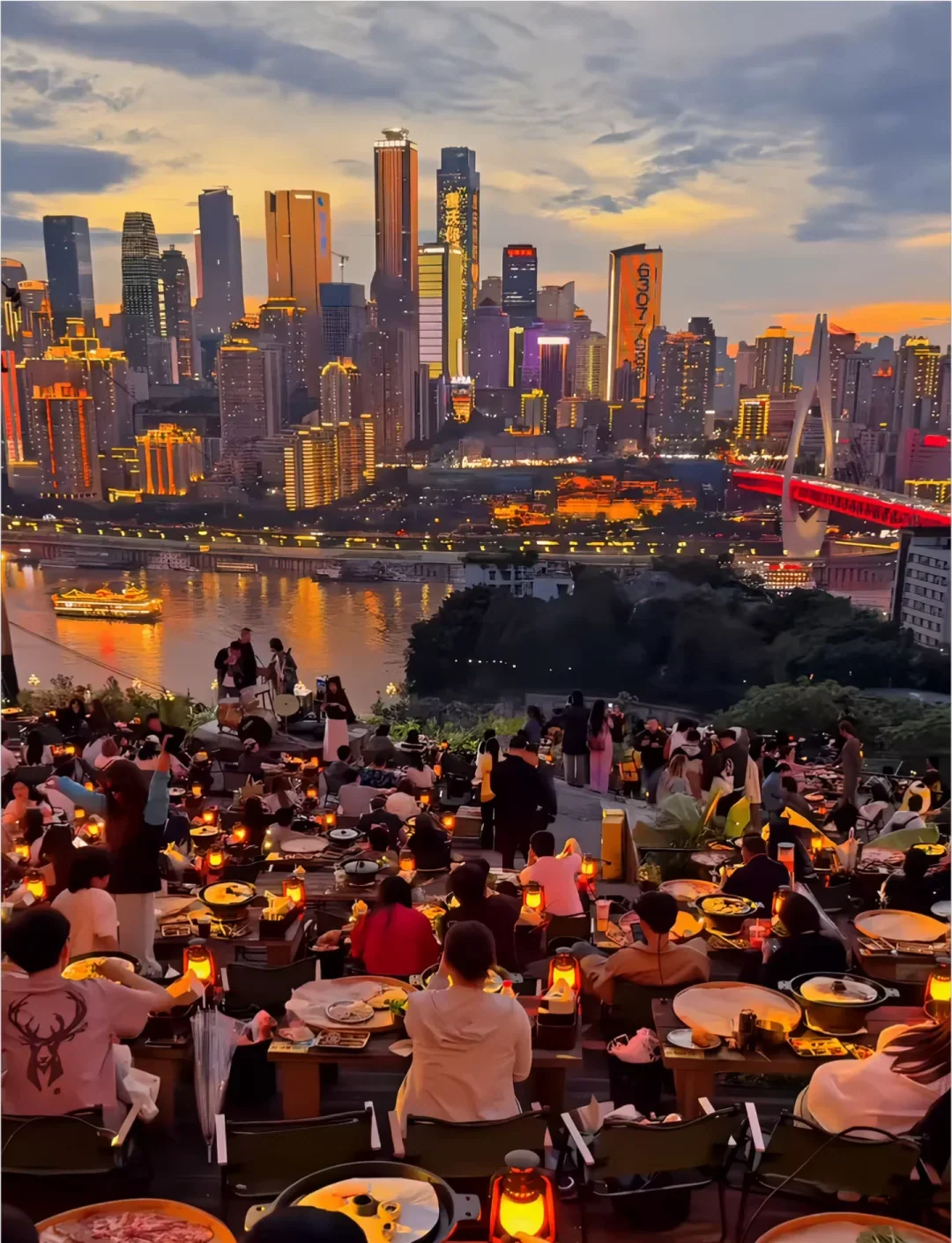 A crowded Chongqing terrace hot-pot dinner facing the river and illuminated Yuzhong skyline at dusk