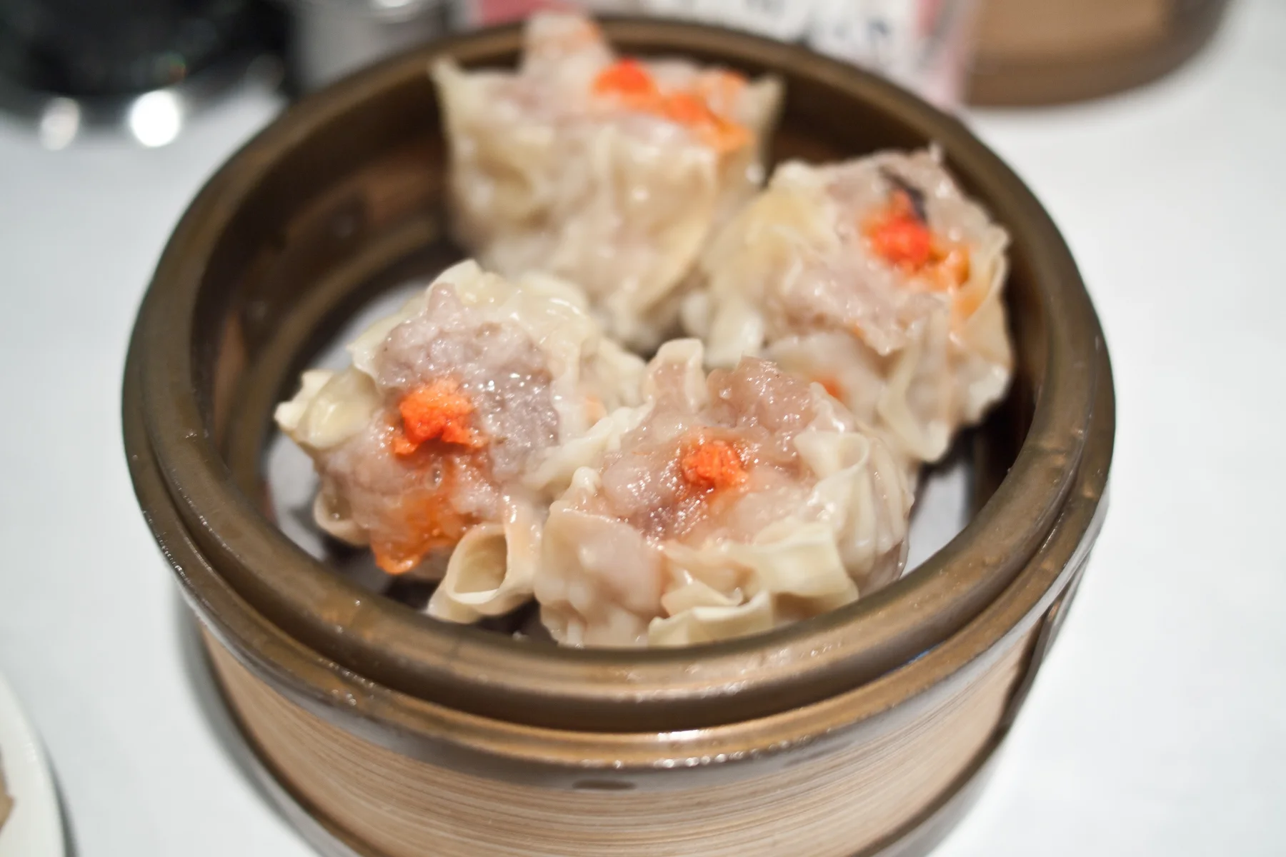Four Cantonese siu mai in a bamboo steamer with gathered wheat wrappers, exposed pork filling and orange topping