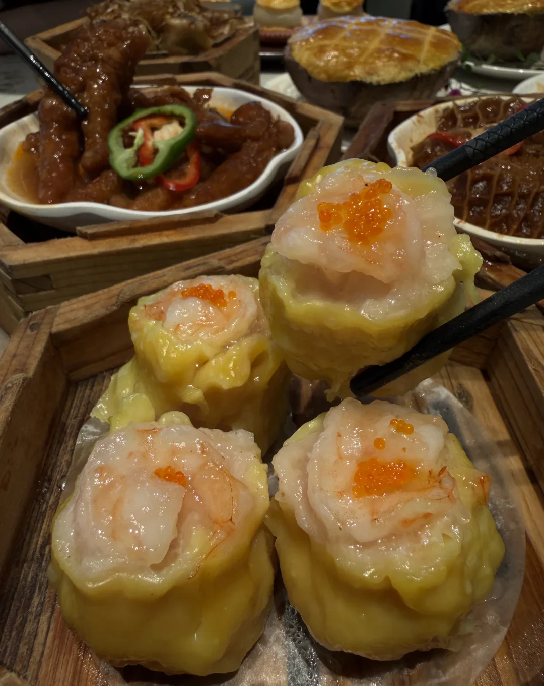 Chopsticks lift one yellow open-topped siu mai above three others in a wooden steamer