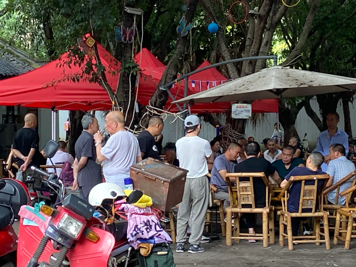 People gathering around bamboo chairs and mahjong tables under trees beside a Chengdu neighborhood teahouse