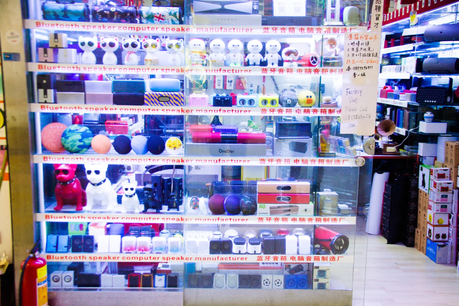 Rows of Bluetooth speakers in different shapes at a Huaqiangbei counter, with a handwritten factory-visit sign, photographed in 2018