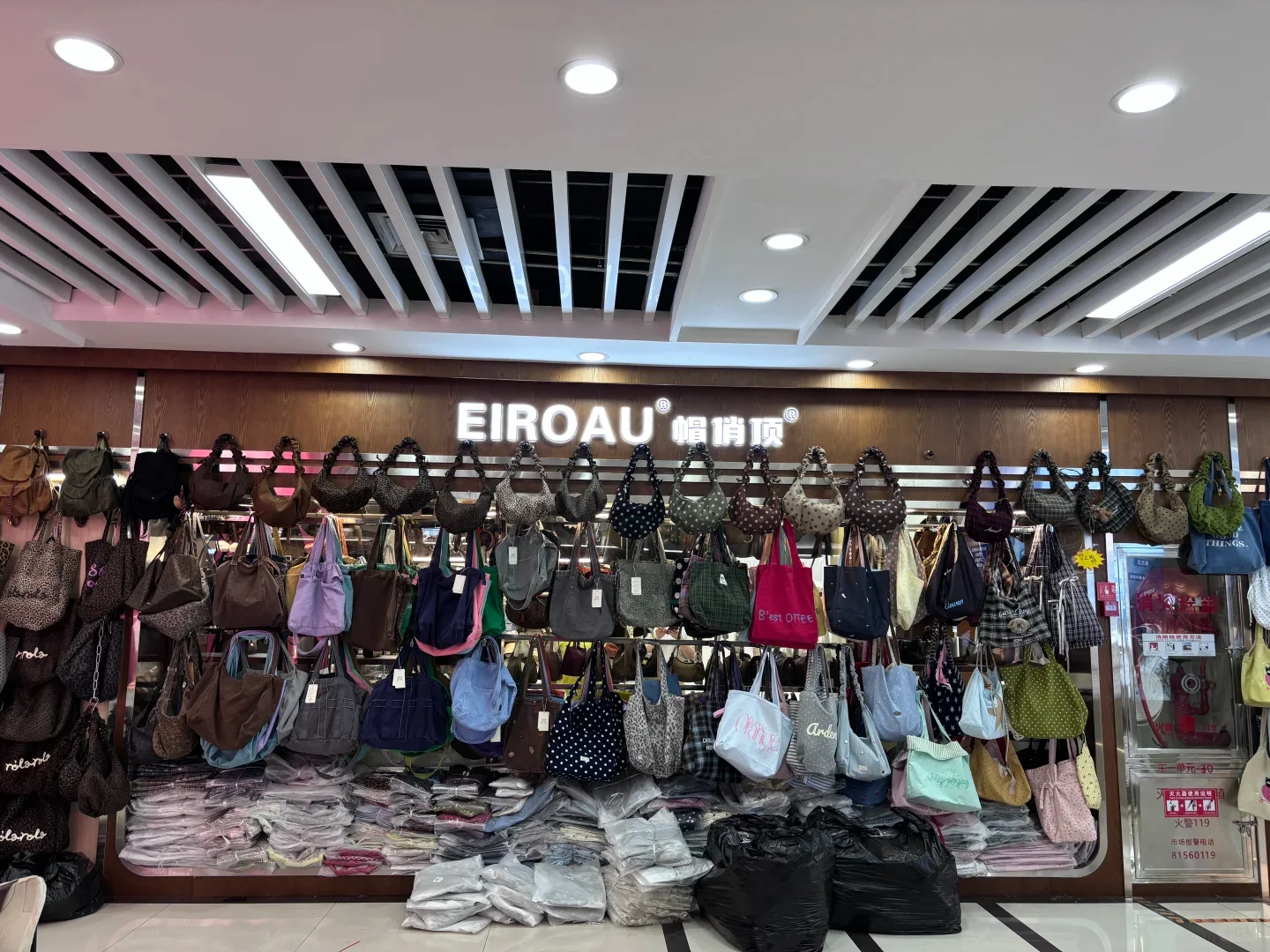Rows of shoulder bags in different colours and shapes hanging above stacks of individually wrapped goods at a Yiwu market shop