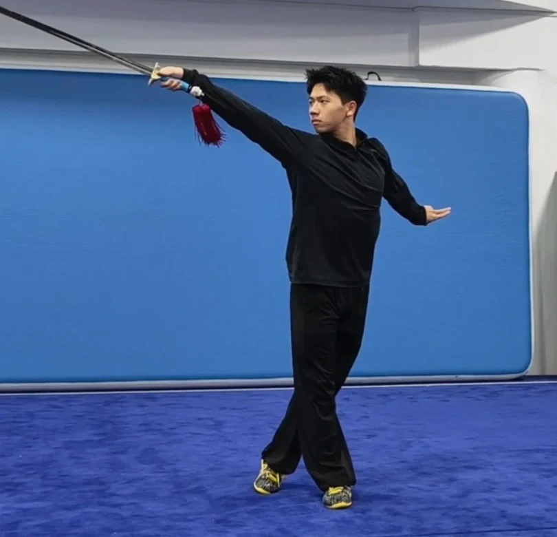 Jianshu practitioner connecting a straight sword line, crossed stance and free hand in one full-body frame