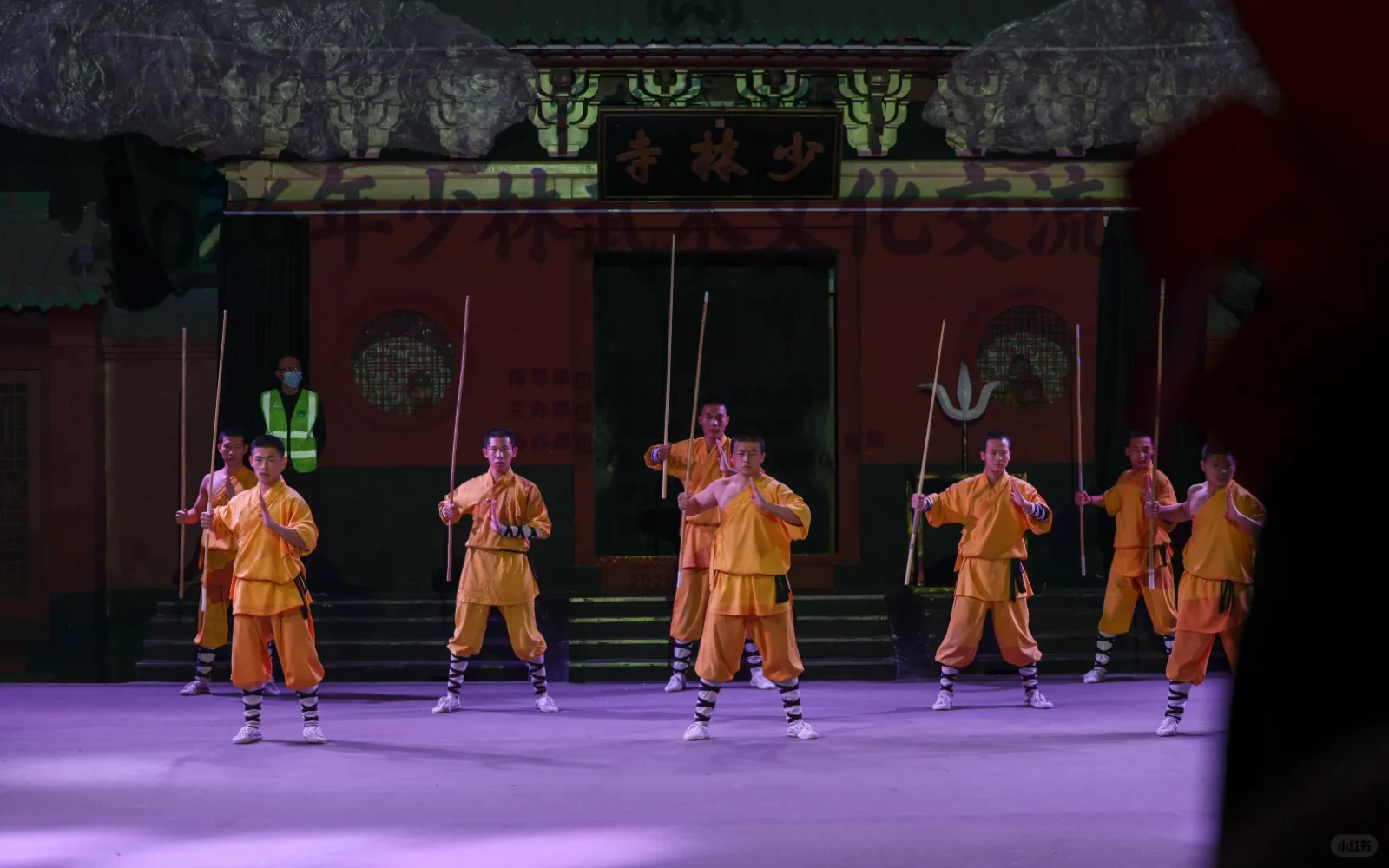 A line of Shaolin-themed stage performers holding staffs and long weapons across a public performance space