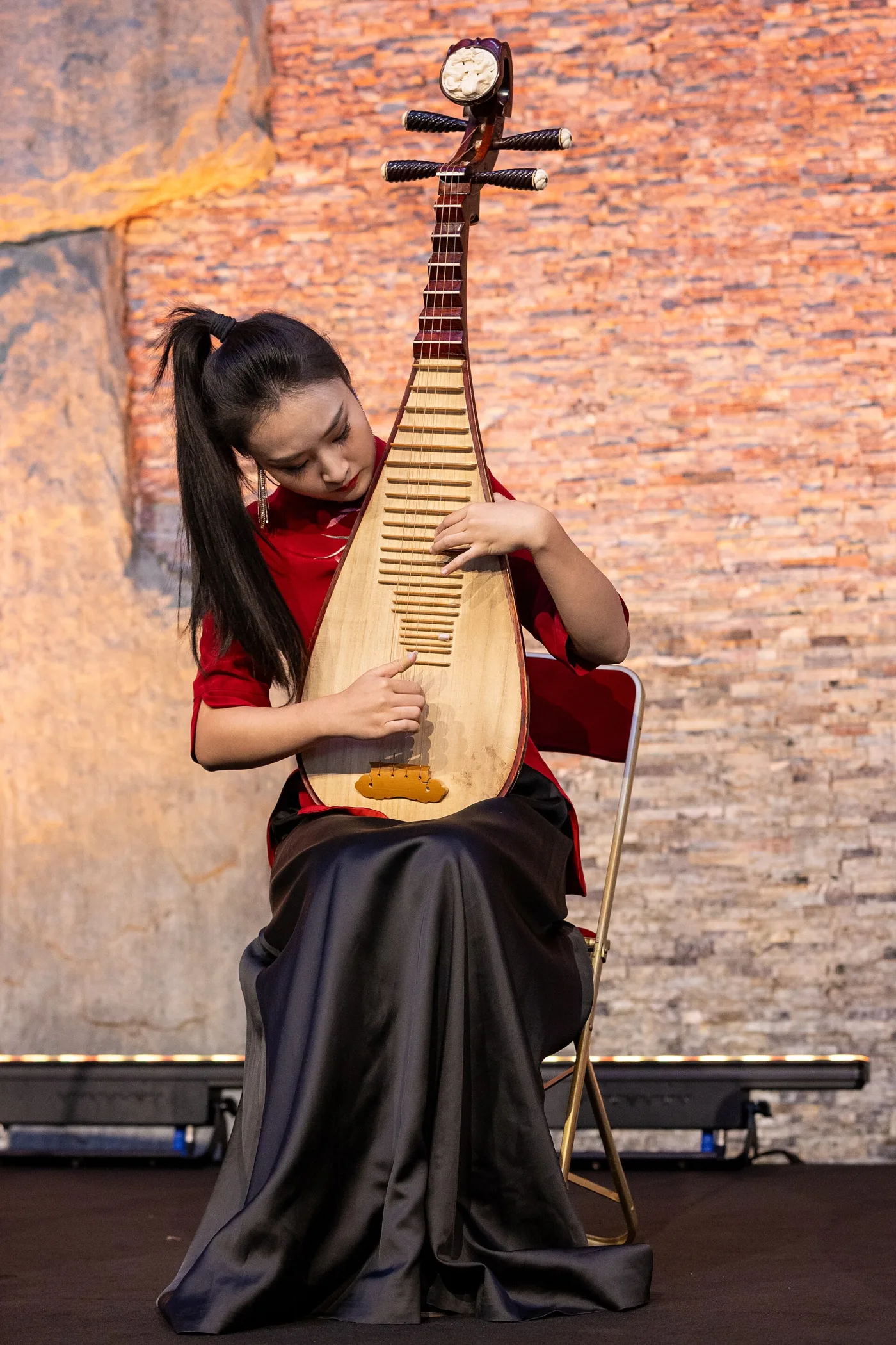 A seated pipa player holds the four-string fretted instrument upright while both hands work across neck and strings