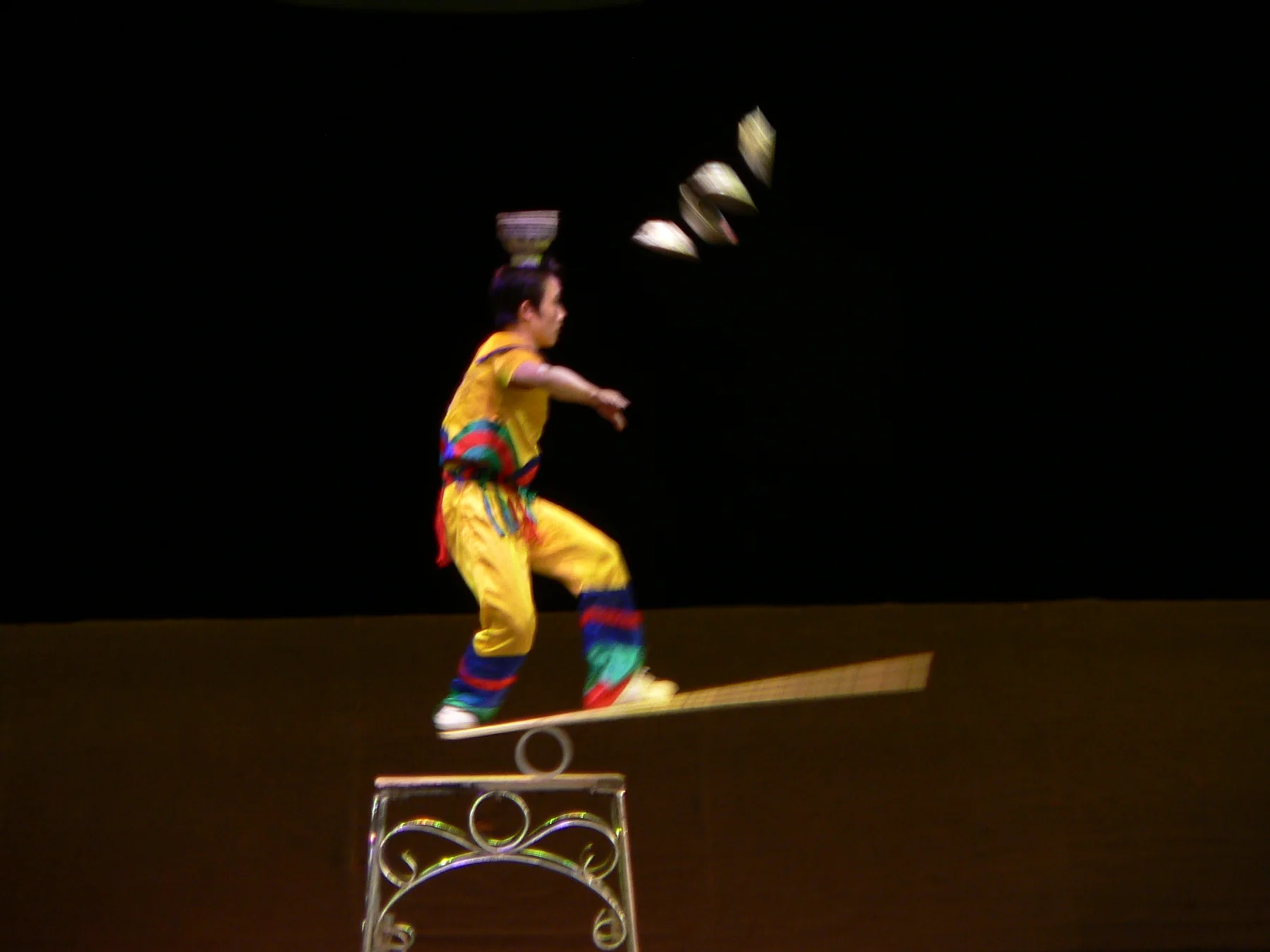 Chinese acrobat airborne from a springboard as several bowls travel toward the receiving performer