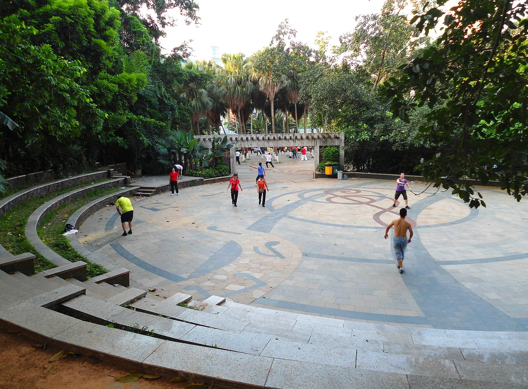 Two adults play badminton across an unmarked open space in Haikou People's Park while other people exercise nearby