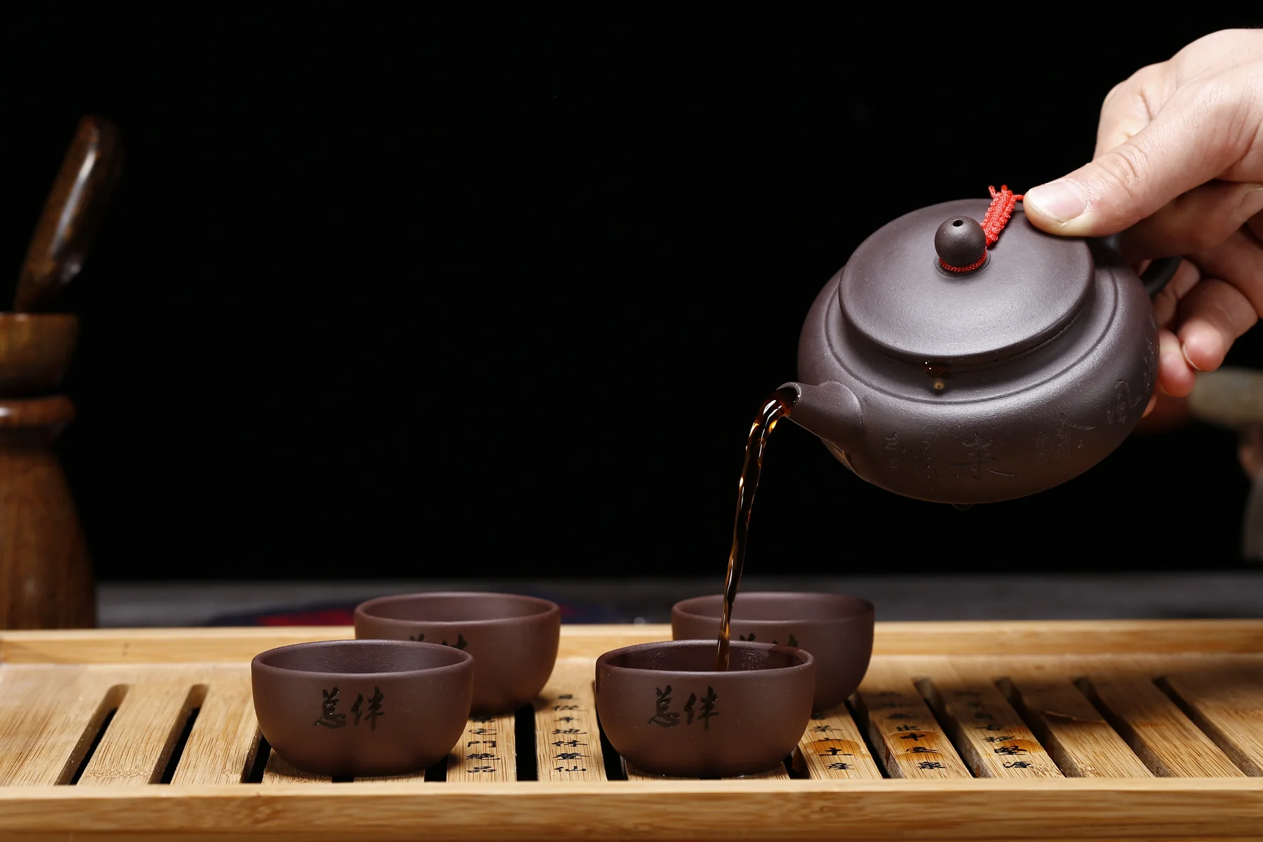 A small dark Chinese teapot pouring tea into matching cups on a bamboo tray