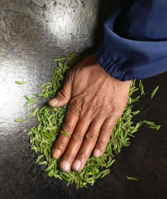 A tea maker pressing and guiding Longjing leaves across a heated wok during shaping