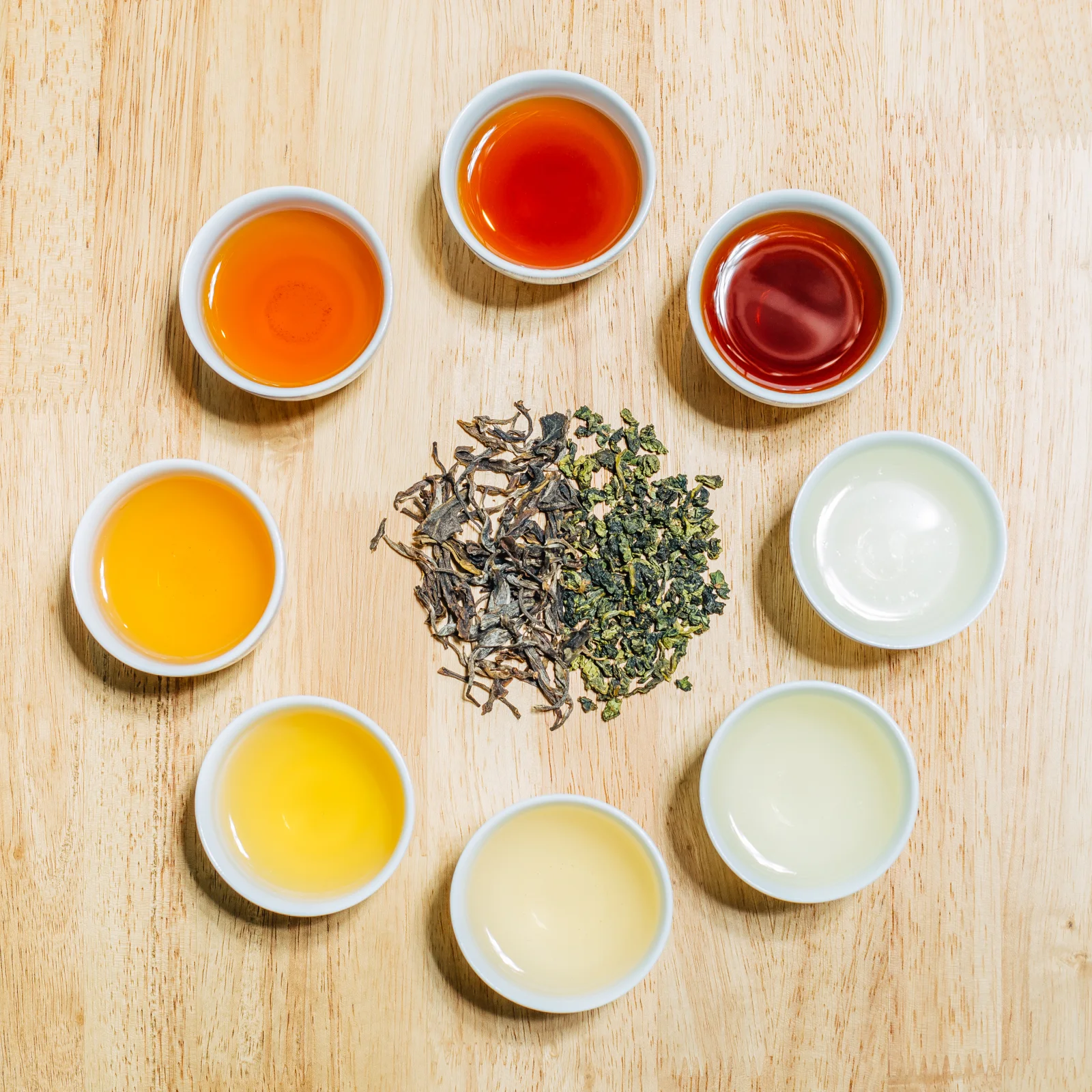 Several pale porcelain Chinese tea cups showing different tea liquor colors beside dry leaves