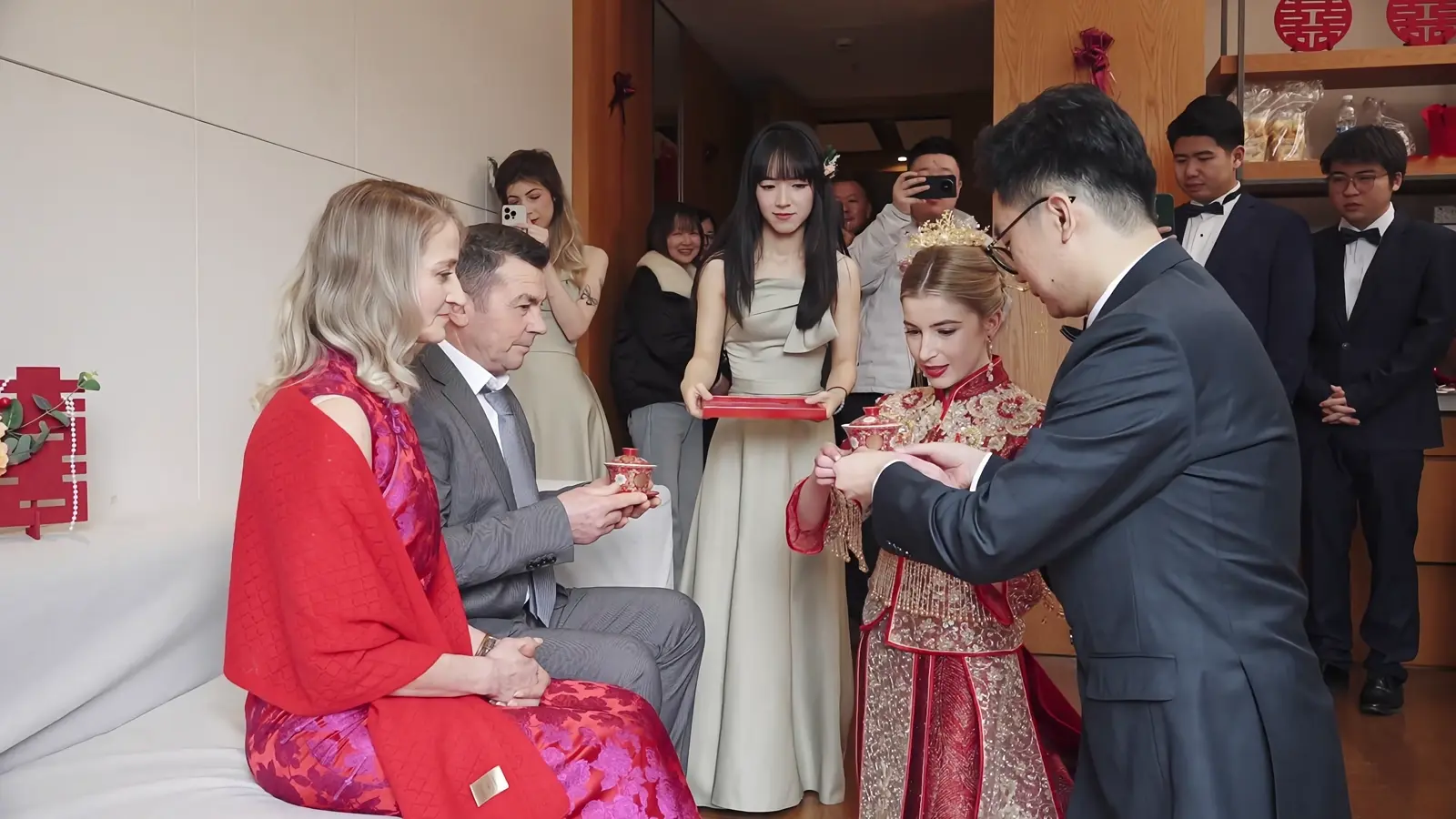 A wedding couple offering covered tea cups to seated parents while relatives watch