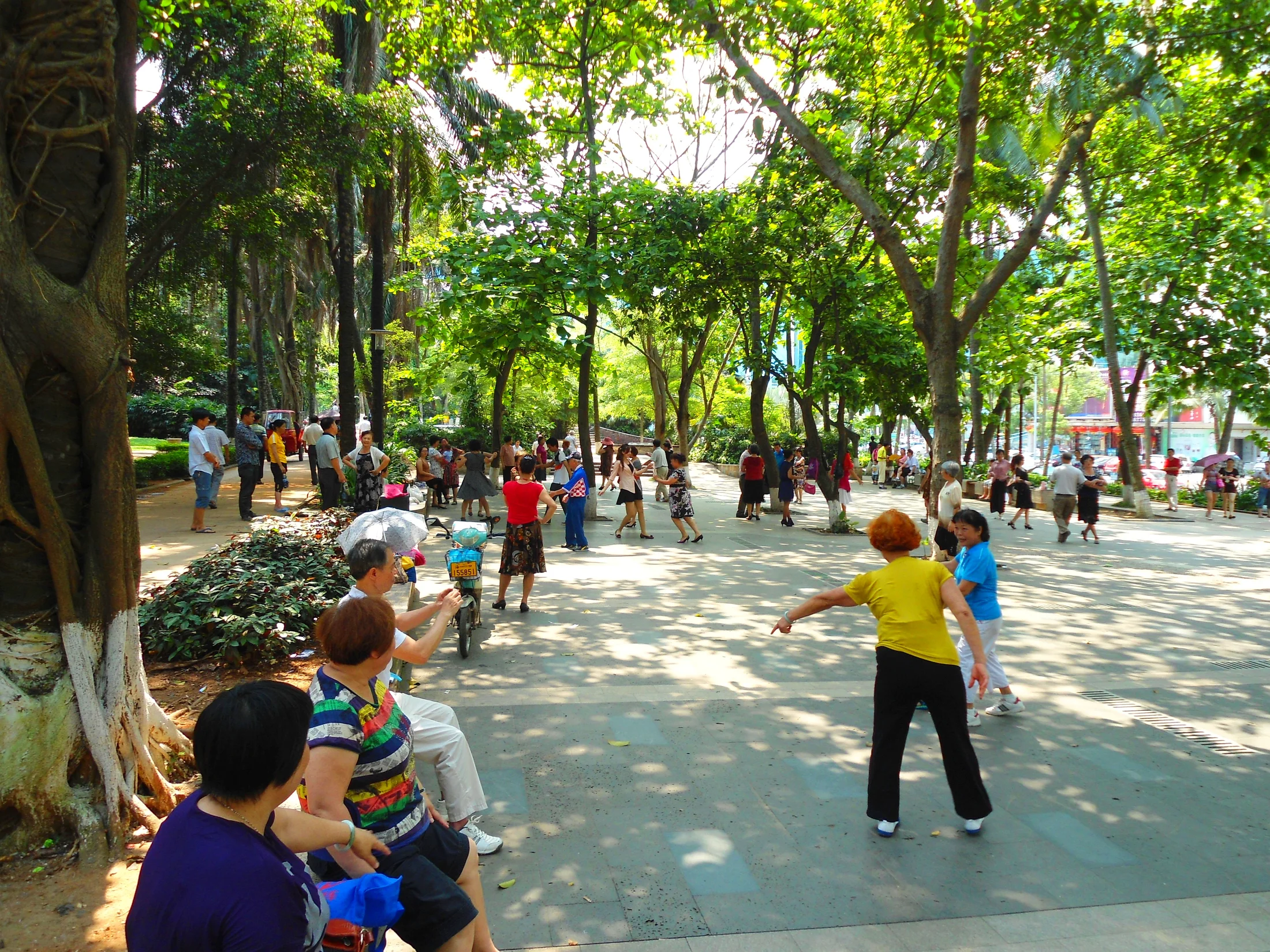 Women dancing and watching along a shaded path in Haikou People's Park