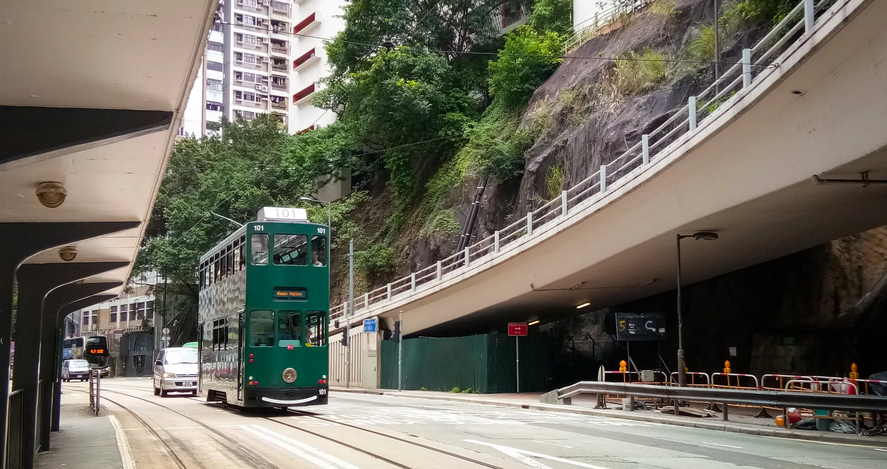 A green Hong Kong tram climbing beside a hillside retaining wall and elevated road