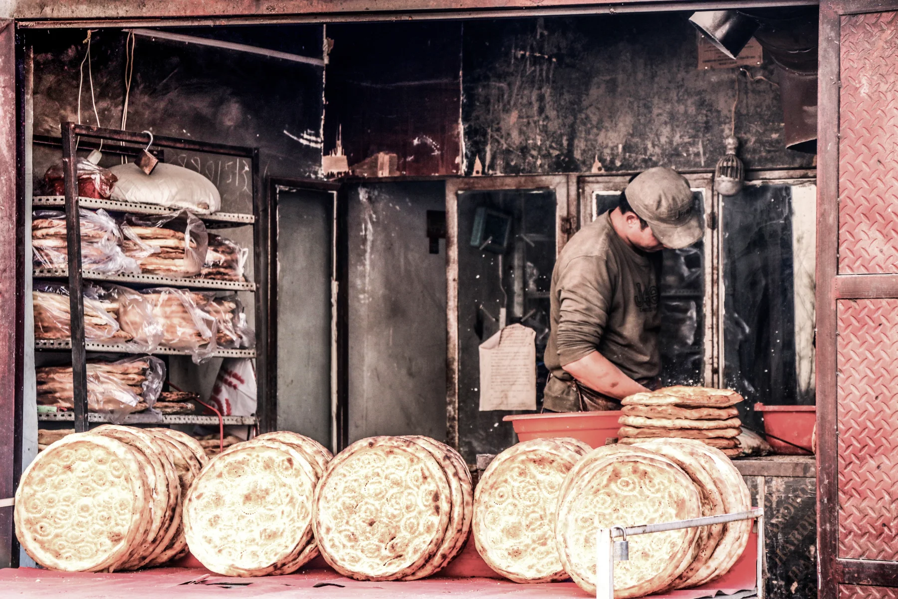 A Kashgar baker preparing large rounds of naan beside a neighborhood oven and bread display