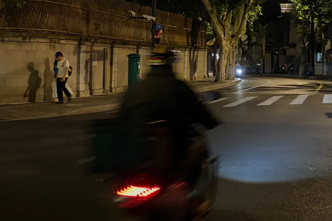 Two pedestrians and a passing scooter on a Shanghai lane after dark, when the city's scale returns to pavement, walls and movement