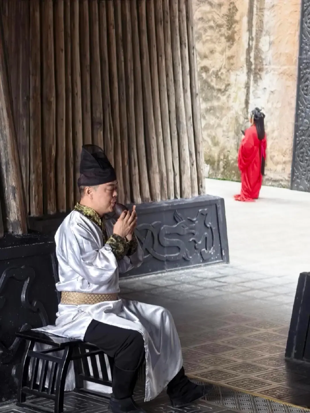 A costumed performer playing in a timber corridor as another figure crosses the bright court beyond