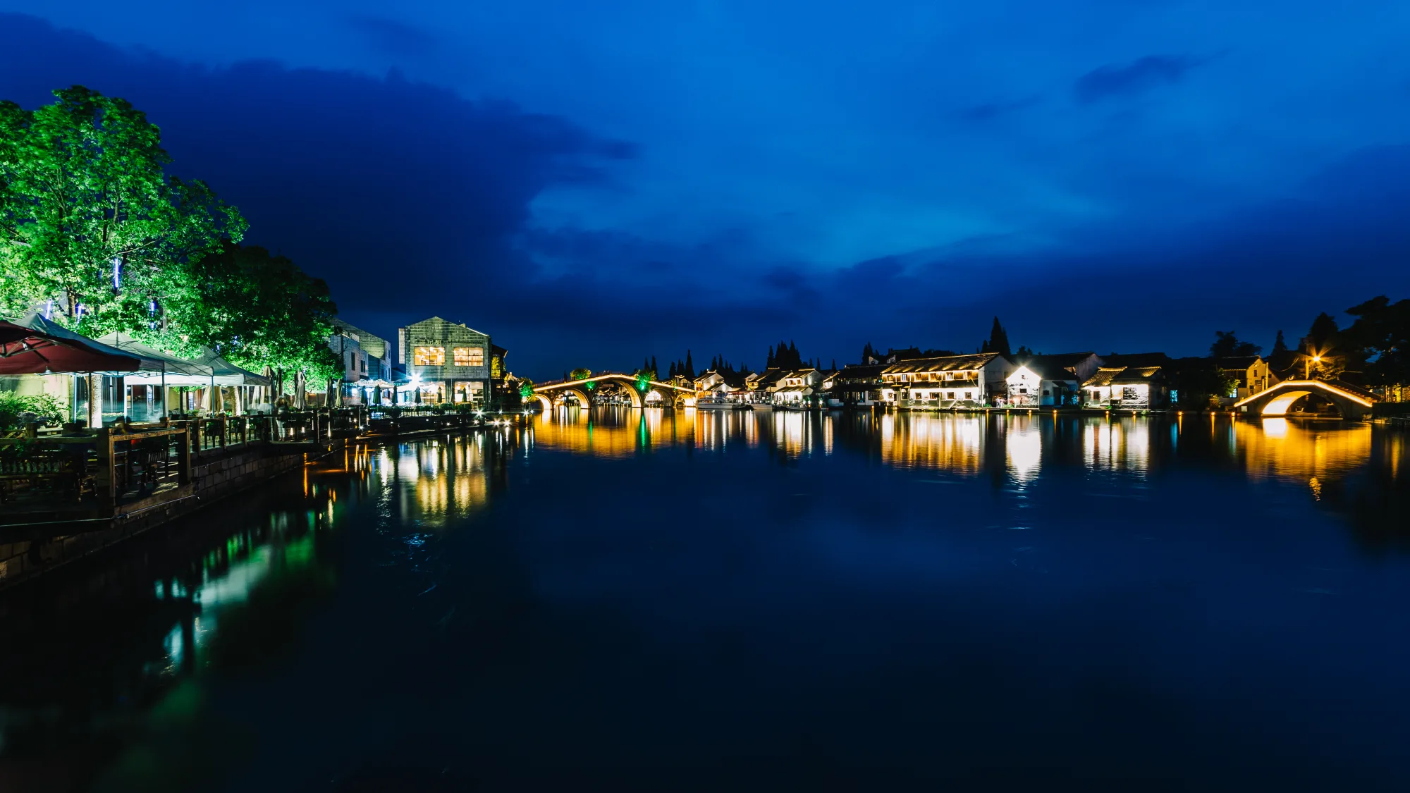 A Jiangnan water town at blue hour with illuminated bridges, white-walled houses and their reflections across the canal
